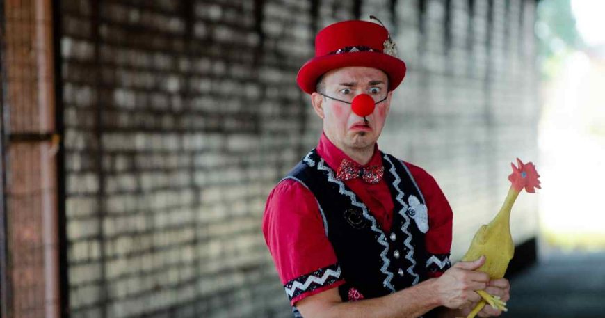 A clown holds a rubber chicken and has a funny frown face.